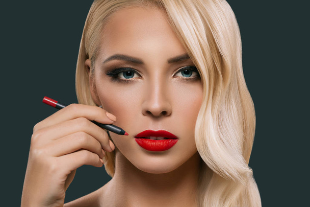 beautiful blonde woman making red lips with cosmetic pencil, isolated on grey - Foto, Bild