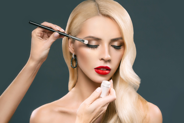 beautiful blonde woman applying glamorous makeup with eyeshadows, isolated on grey - Photo, Image