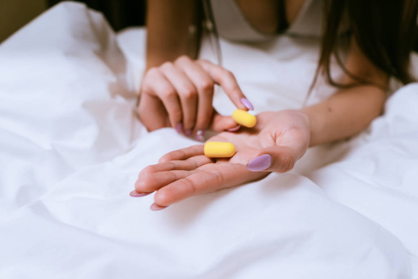 girl sitting on a bed under a white blanket, on her arm yellow earplugs against street noise - Photo, Image