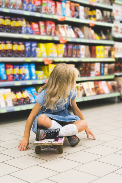 blonde kid sitting on skateboard in supermarket with shelves behind - Photo, Image