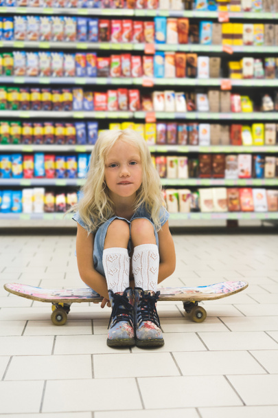adorable blonde child sitting on skateboard in supermarket with shelves behind - Fotó, kép