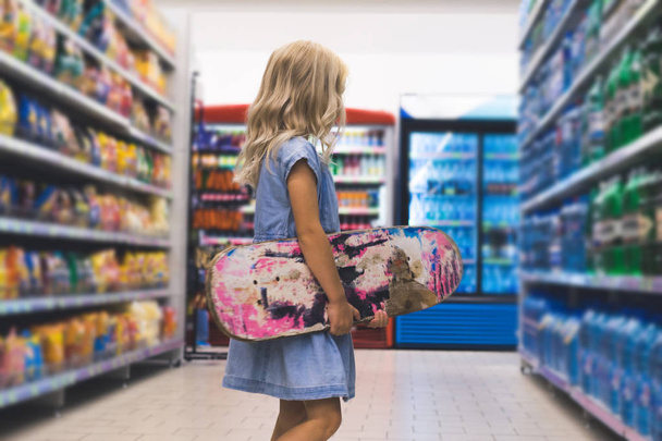 blonde kid with skateboard standing in supermarket with shelves behind - Foto, afbeelding