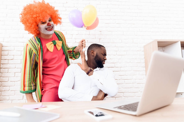 A man in a clown suit is standing beside a black man who sits at his desk. - Photo, image