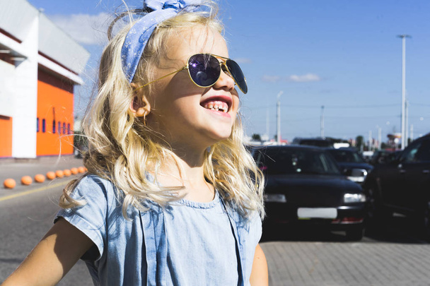 стильна весела чарівна дитина в сонцезахисних окулярах стоїть на парковці з машинами
 - Фото, зображення