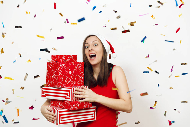 Pretty surprised young happy woman dressed in red dress and Christmas hat holding gift boxes on white background and colorful confetti. Santa girl with present isolated. New Year holiday 2018 concept - Фото, изображение