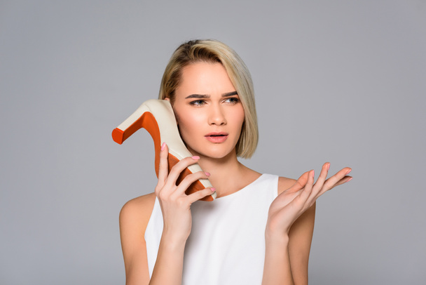 beautiful confused girl holding heeled shoe as phone, isolated on grey - Photo, Image