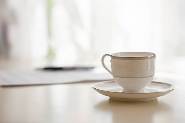 Closeup of white coffee cup with plate standing on table with documents and pen, blurred background - Fotó, kép