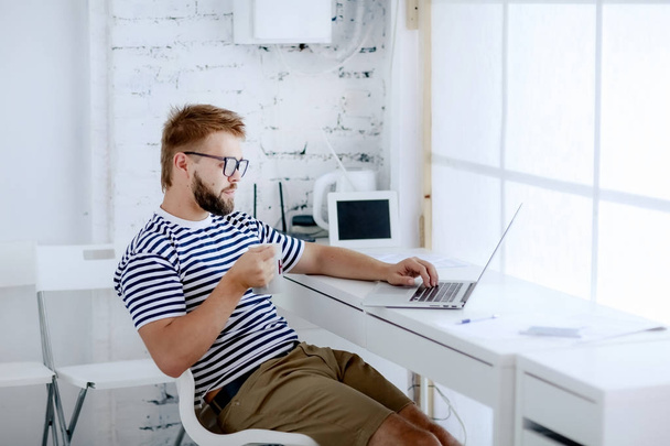 young handsome businessman working on laptop sitting at desk and drinking coffee in office - 写真・画像