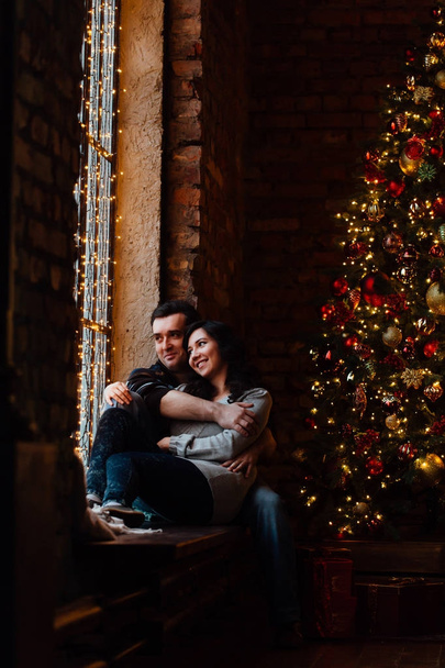 Couple of lovers hugs sitting on the windowsill in the Christmas loft studio. guy is hugging the girl. - Фото, изображение