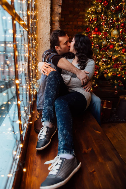 Couple of lovers hugs sitting on the windowsill in the Christmas loft studio. guy is hugging the girl. - 写真・画像