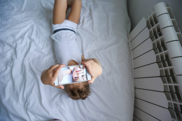 view from above on little boy lying on white bed taking selfie with smartphone - Fotoğraf, Görsel