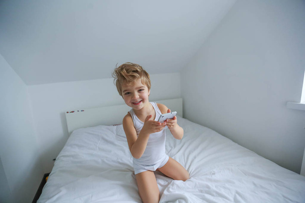 view from above on little boy sitting on white bed playing on smartphone - Fotografie, Obrázek