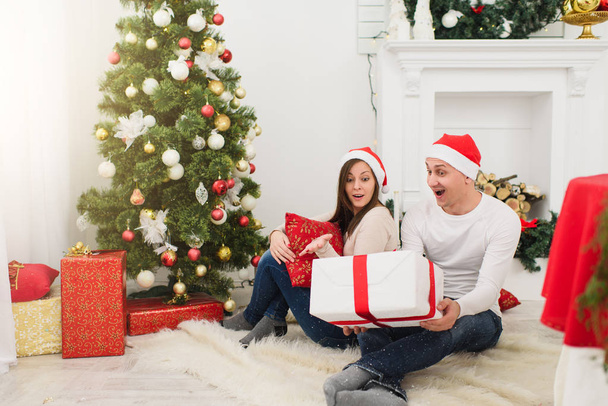 Mutlu neşeli komik genç çift dekore edilmiş yeni yıl ağacı ve hediye kutuları arka planda Noel çelenk ışık Oda evde oturan kırmızı şapkalı aşık. Aile, tatil 2018 kavramı. - Fotoğraf, Görsel