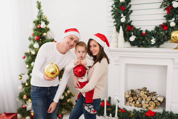 Happy cheerful parents with cute little son. Child boy with big red tree toy ball standing in decorated New Year light room at home on the background of Christmas wreath. Family, holiday 2018 concept. - Fotoğraf, Görsel