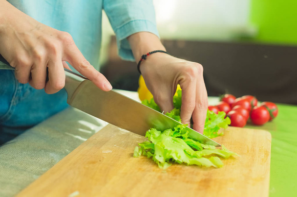 Hands of a young girl slice green lettuce leaves on a wooden cutting board on a green table in a home setting against a background of red cherry tomatoes. - 写真・画像