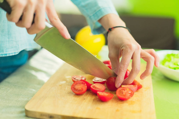 Hands of a young girl chop the cherry tomatoes on a wooden cutting board on a green table in a home setting - Foto, Imagem
