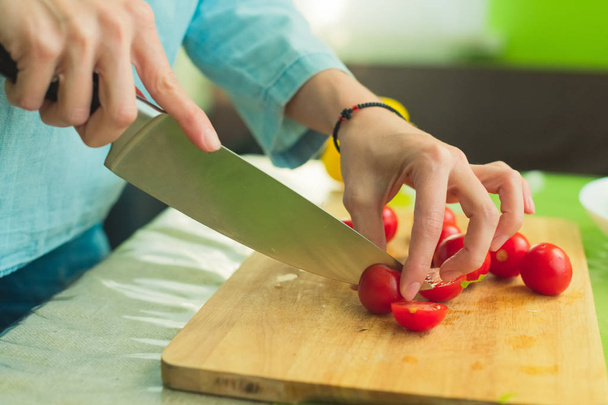 Hands of a young girl chop the cherry tomatoes on a wooden cutting board on a green table in a home setting - Photo, Image