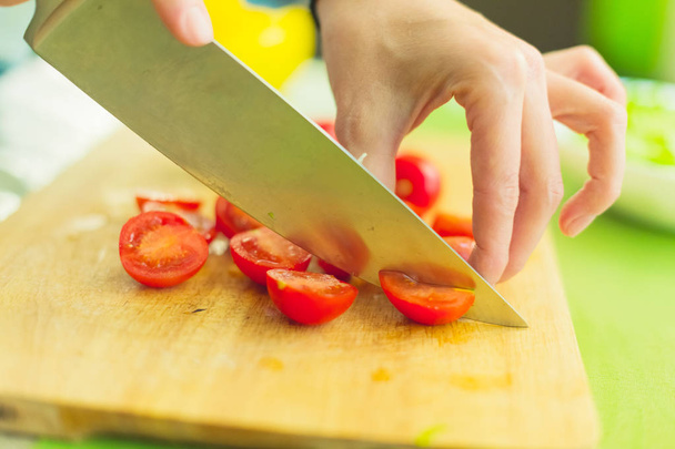 Hands of a young girl chop the cherry tomatoes on a wooden cutting board on a green table in a home setting - Photo, Image