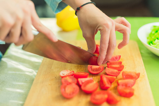 Hands of a young girl chop the cherry tomatoes on a wooden cutting board on a green table in a home setting - Foto, Bild