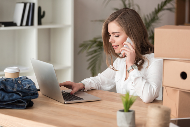 Zijaanzicht van jonge zakenvrouw praten op smartphone tijdens het werk op laptop - Foto, afbeelding