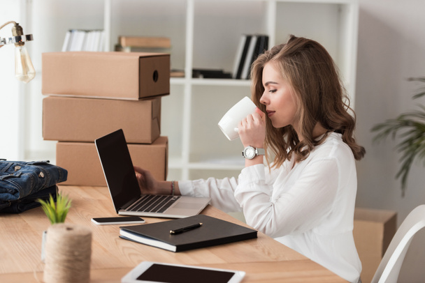 vista lateral de empresária beber café enquanto trabalhava no laptop à mesa
 - Foto, Imagem