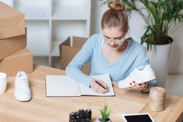 portrait of young entrepreneur with pair of shoes making notes in notebook at home office - Photo, Image