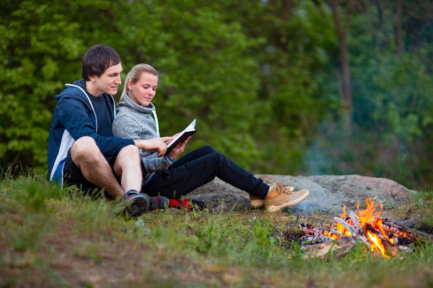 A couple of tourists in time of the hike, sitting and relaxing near the fire embracing, The woman read a book, The guy hugs the girl. The concept of love, tenderness and recreation. Shallow depth of field, selective focus - Photo, Image