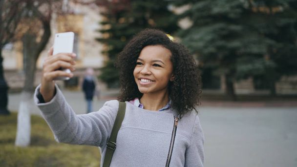 Cheerful mixed race student girl talking at video call with smartphone near univercity - 写真・画像