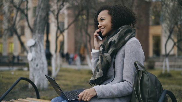 Attractive african american student girl talking her phone sitting on bench with laptop computer outdoors - Фото, изображение