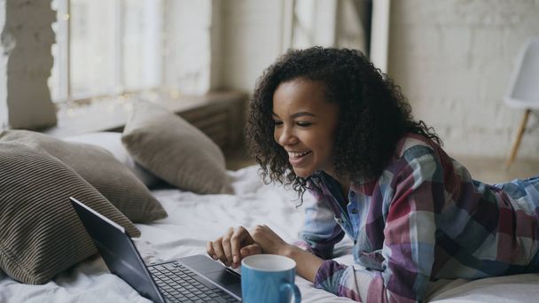 Curly african american girl lauging using laptop for sharing social media lying in bed at home - Foto, Bild