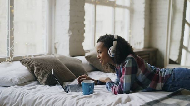 Attractive mixed race girl listen to music while surfing social media on laptop lying on bed - Photo, Image