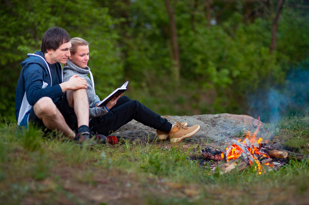 Młoda Para turystów pieszych, siedzącej i odpoczynku w pobliżu ognia obejmując, Kobieta czytać książki, facet przytula dziewczynę. Pojęcie miłości, czułości i rekreacji. Płytkiej głębi ostrości pola, selektywne - Zdjęcie, obraz
