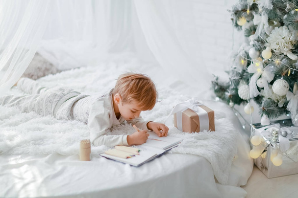 little boy lying on white bed near Christmas tree and writing in notebook - Foto, Bild