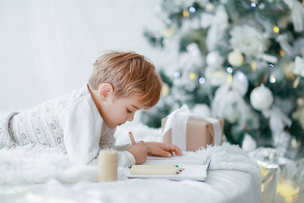 little boy lying on white sofa under Christmas tree and making wish list - Foto, afbeelding