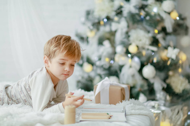 little boy lying on white bed near Christmas tree and writing in notebook - Foto, afbeelding