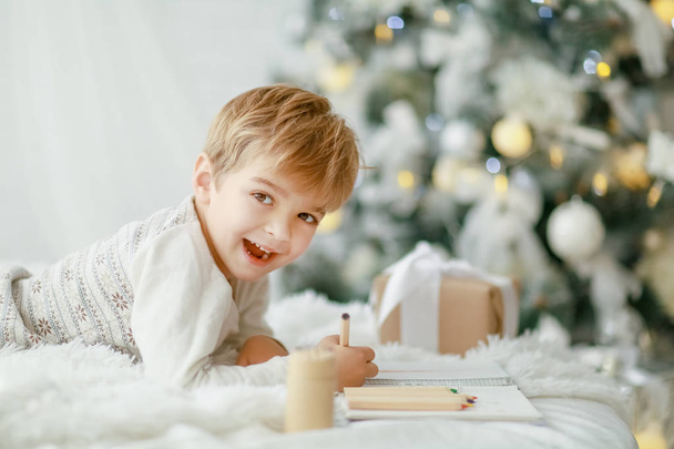 little boy lying on white sofa under Christmas tree and making wish list - Foto, Bild