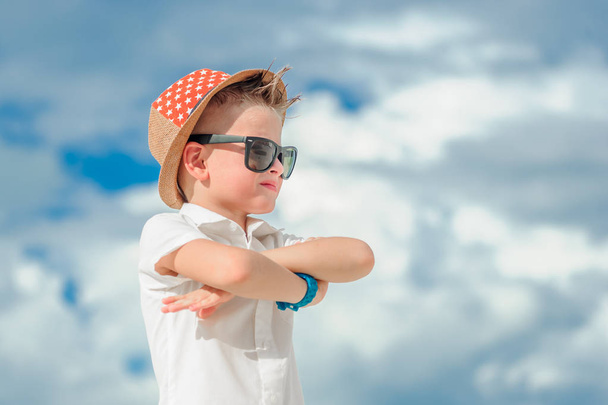 A close up portrait on a background of clouds: handsome little boy dressed in solid bright shirt with sunglasses and a fedora hat. He crossed his arms, turned sideways, looking far away - Фото, изображение