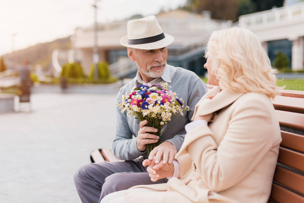 Two pensioners are sitting on a bench in the alley. An elderly man gives a woman flowers - Foto, Imagen
