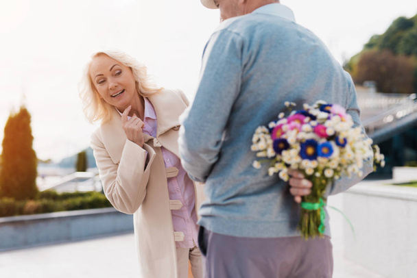 An elderly man is holding a bouquet of flowers behind back. A woman came on a date and tries to spy on what hides a man - Foto, imagen