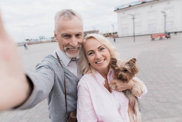 An elderly couple makes selfie on a beautiful square. A woman is holding a small dog in her hands - Fotó, kép