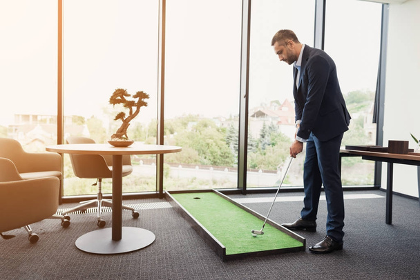 A man in a strict business suit plays in an office in a mini-golf - Foto, afbeelding