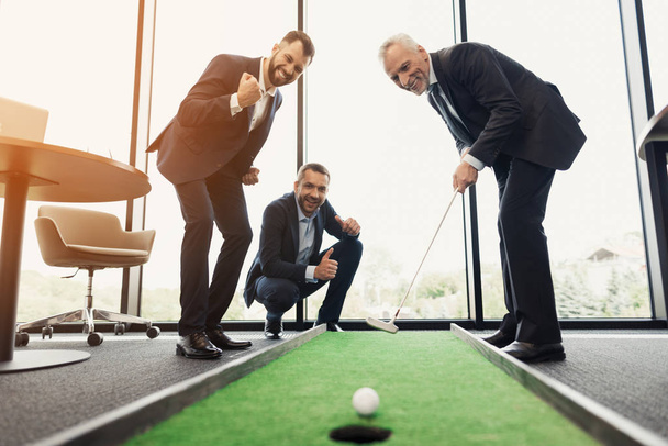 A respectable elderly man is playing golf in his office. Next to him are his workers. They look like the boss is playing - Foto, imagen