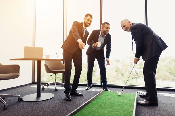 A respectable elderly man is playing golf in his office. Next to him are his workers. They look like the boss is playing - Zdjęcie, obraz