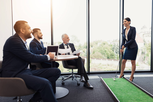 Three men sit at a table and watch a woman in a business suit playing mini golf in the office - Photo, Image