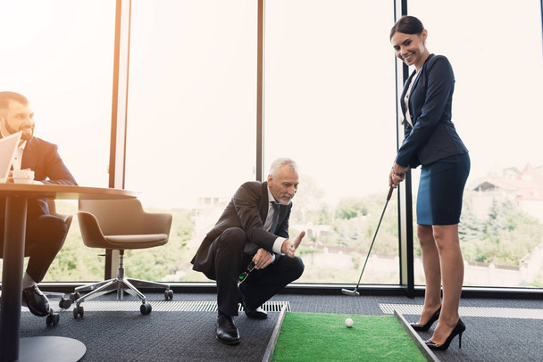 A woman in a black business suit plays golf in the office. An old man in a business suit helps her - Photo, Image