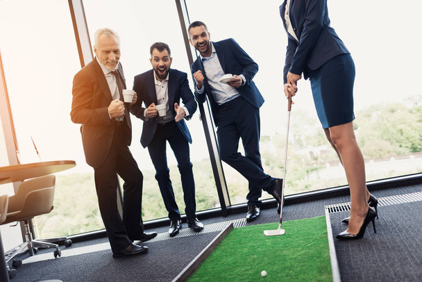 Три бизнесмена и деловые леди играют в гольф. Женщина держит в руках клюшку для гольфа. Мужчины смотрят
 - Фото, изображение
