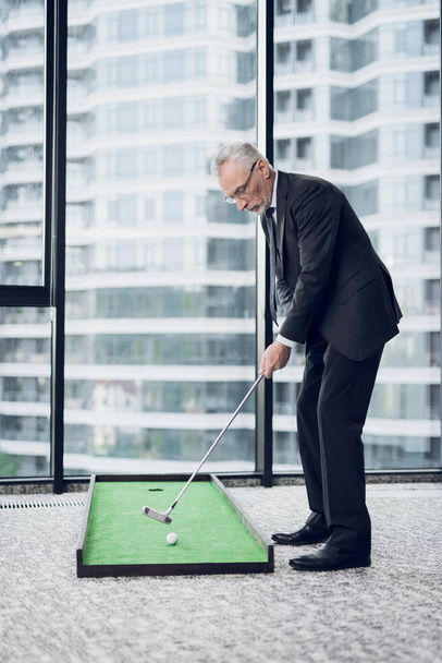 Un respetable anciano jugando un mini golf en la oficina. Se está preparando para atacar.
 - Foto, Imagen