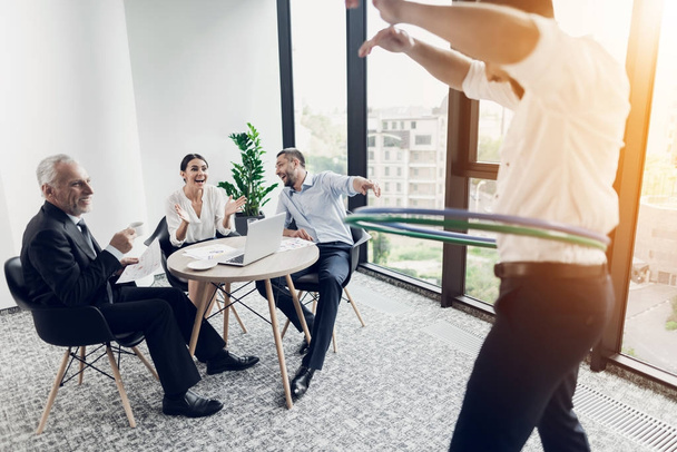 Businessman in the office turns hula-hoop. His colleagues sit at the table and look at him. They are very fun - Foto, afbeelding