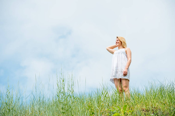 Mooie vrouw in korte witte jurk, poseren en lachend op een weide op warme zonnige dag. Blauwe hemel en gras achtergrond. - Foto, afbeelding