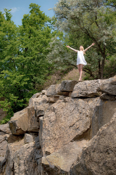 De jonge vrouw met opgeheven handen sky op de rand van de klif in zomerdag. Meisje dragen korte witte jurk.  - Foto, afbeelding